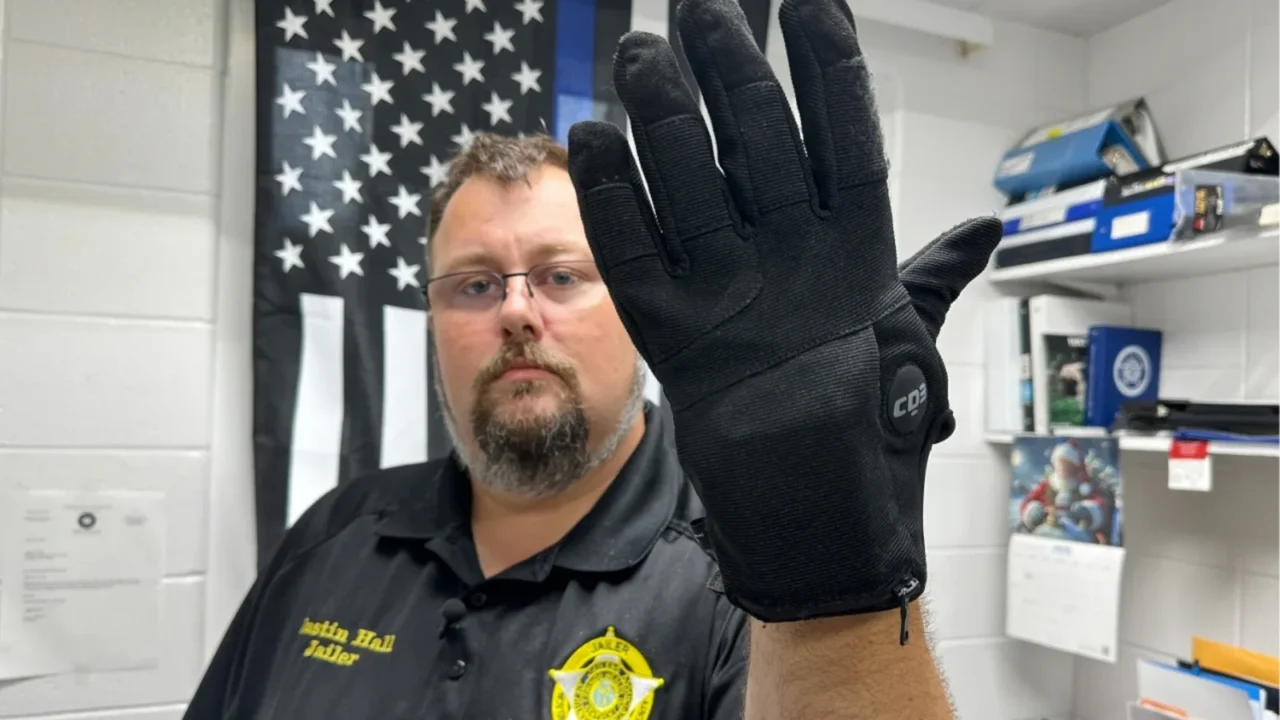 Policial exibe luva tátil de contenção em frente a bandeira americana, em ambiente de trabalho com prateleiras e equipamentos ao fundo