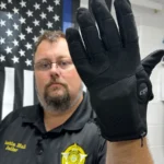 Policial exibe luva tátil de contenção em frente a bandeira americana, em ambiente de trabalho com prateleiras e equipamentos ao fundo