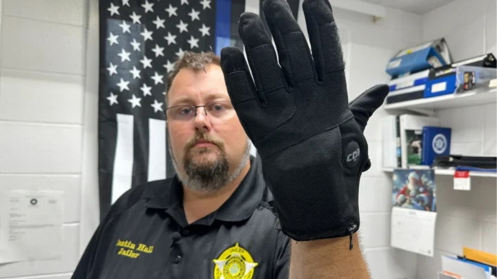 Policial exibe luva tátil de contenção em frente a bandeira americana, em ambiente de trabalho com prateleiras e equipamentos ao fundo