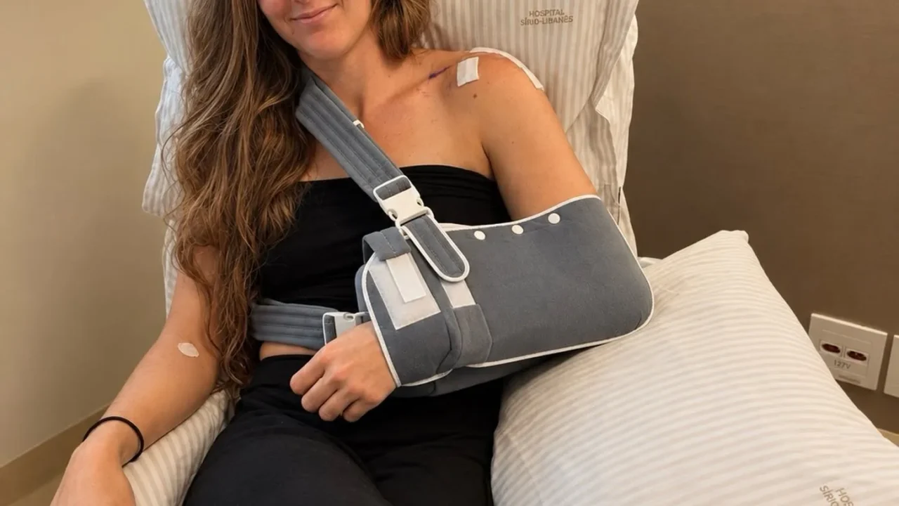 Mulher com braço esquerdo imobilizado em tipoia posa em cama de hospital após cirurgia no ombro