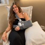 Mulher com braço esquerdo imobilizado em tipoia posa em cama de hospital após cirurgia no ombro