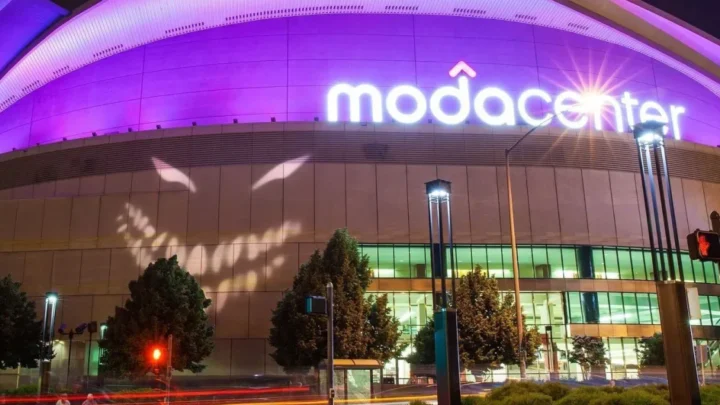 Fachada iluminada do Moda Center, arena coberta com estrutura de vidro e metal, com o nome em letras luminosas roxas na parte superior e vegetação ao entorno durante a noite