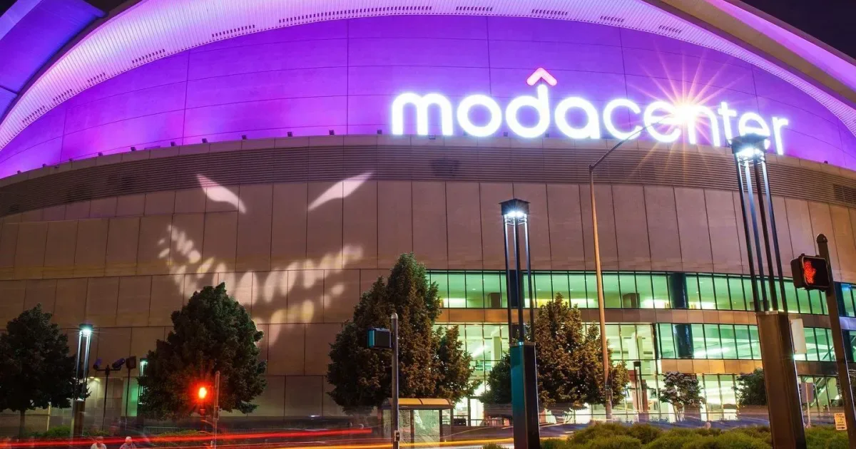 Fachada iluminada do Moda Center, arena coberta com estrutura de vidro e metal, com o nome em letras luminosas roxas na parte superior e vegetação ao entorno durante a noite
