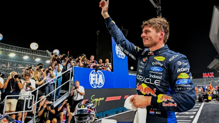 Piloto da Red Bull com macacão azul acena para fotógrafos em pista noturna, com capacete e toalha sobre a bancada à frente