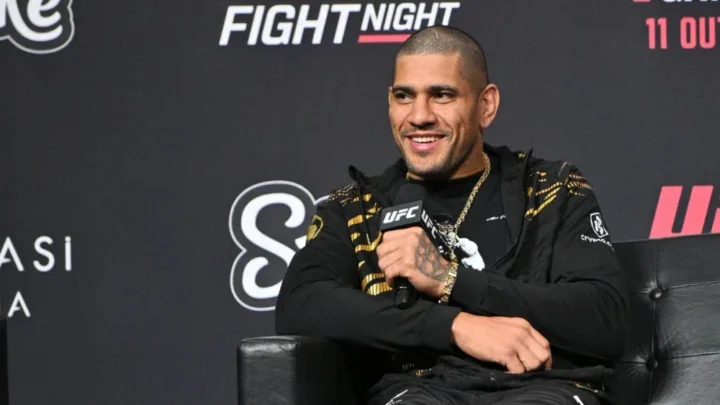 Homem de camisa preta com detalhes dourados sorri enquanto segura um microfone da UFC, sentado em poltrona de couro diante de painel do evento UFC Fight Night