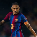 Jogador de camisa listrada em azul e grená, com braçadeira do Barcelona, olha em direção ao campo durante uma partida noturna