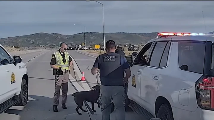 O policial Wayne Henderson, de colete, ao lado do cão Bruno, um labrador chocolate, entre viaturas da Patrulha Rodoviária de Utah no viaduto interditado
