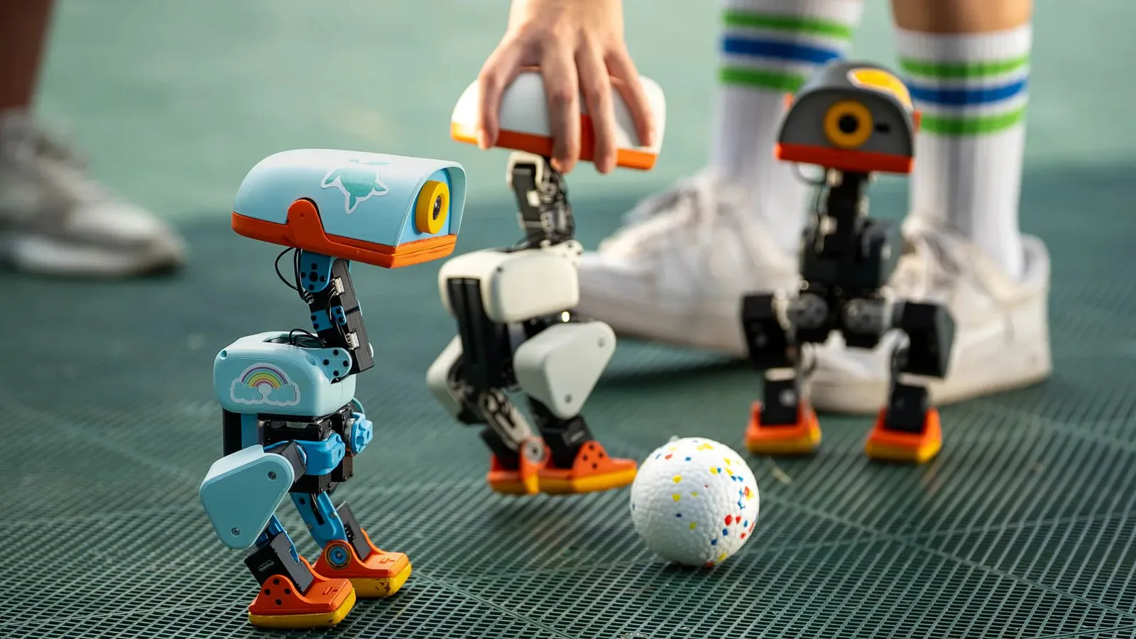 Três robôs Microduck, um azul, um branco e um preto, ao redor de uma bola sobre um piso verde, enquanto uma mão segura um deles