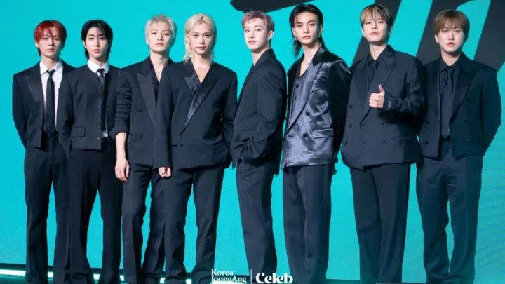 Oito integrantes de um grupo de K-pop posam em fila usando ternos pretos contra fundo azul turquesa em evento de mídia