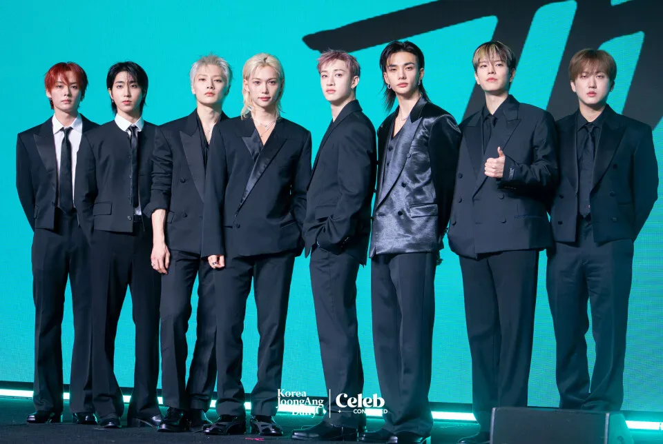 Oito integrantes de um grupo de K-pop posam em fila usando ternos pretos contra fundo azul turquesa em evento de mídia