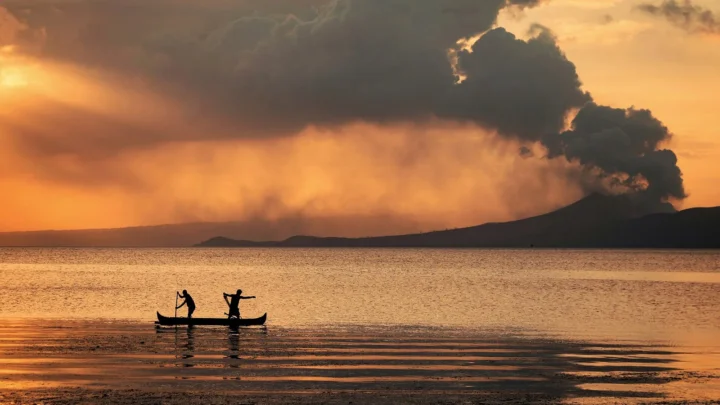 Coluna de cinzas do vulcão Taal sob céu alaranjado, vista do lago, com dois homens num barco em silhueta, dias depois da erupção de janeiro de 2020