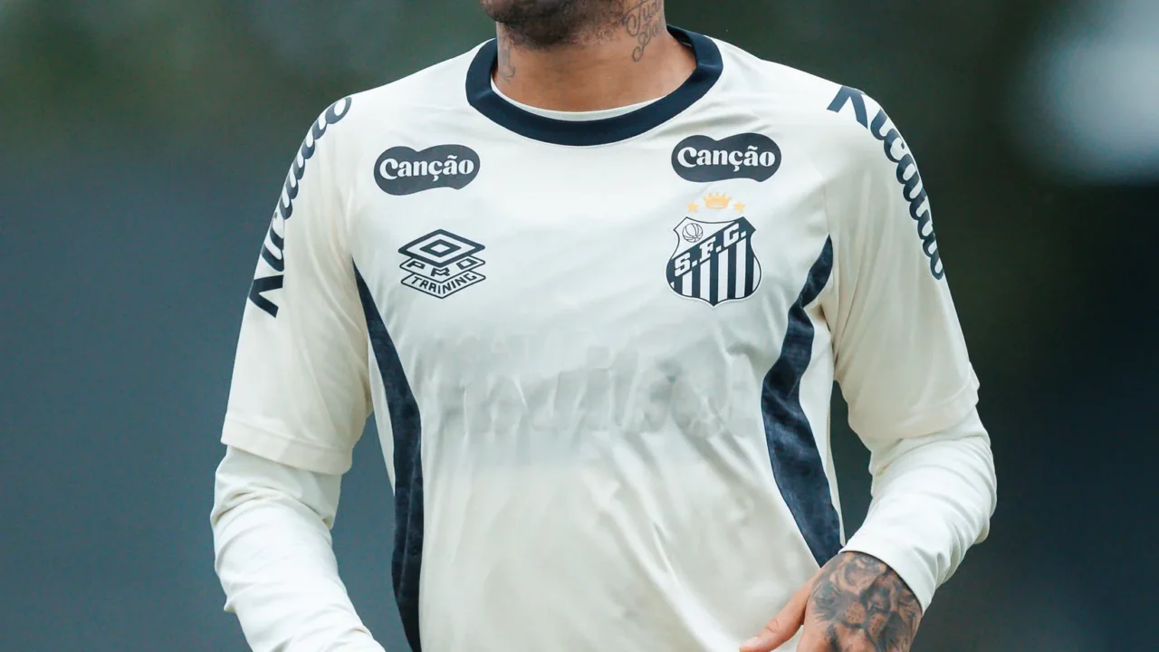Jogador de camisa branca com detalhes pretos e escudo do Santos, com tatuagens nos braços, em treino em campo de grama