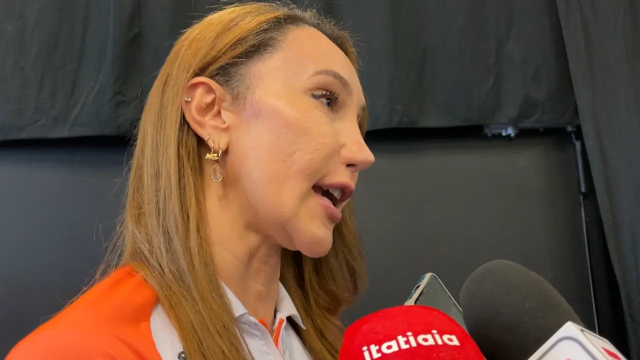 Mulher de cabelo castanho claro, usando blazer laranja e camisa branca, fala ao microfone da Rádio Itatiaia durante entrevista