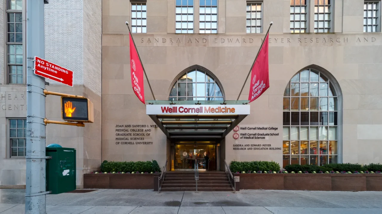 Fachada da Weill Cornell Medicine na York Avenue, em Manhattan, com a entrada principal e duas bandeiras vermelhas com o nome da instituição