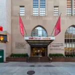 Fachada da Weill Cornell Medicine na York Avenue, em Manhattan, com a entrada principal e duas bandeiras vermelhas com o nome da instituição