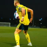 Jogador de camisa amarela com faixa de capitão e número 7 nas costas comemora em campo gramado à noite, com a lateral do uniforme visível