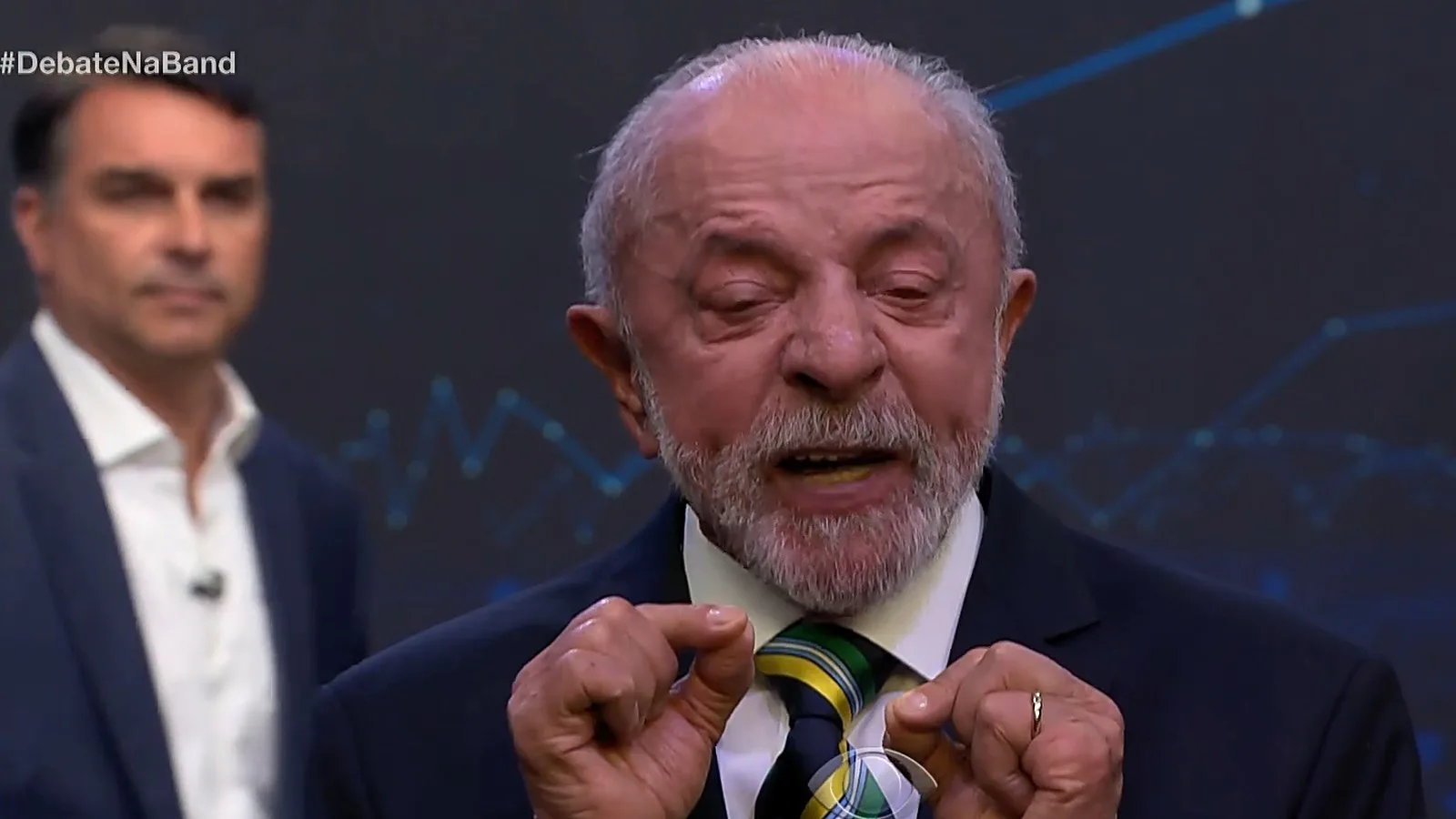 Lula fala em primeiro plano, com Flávio Bolsonaro desfocado ao fundo, durante o 2º bloco