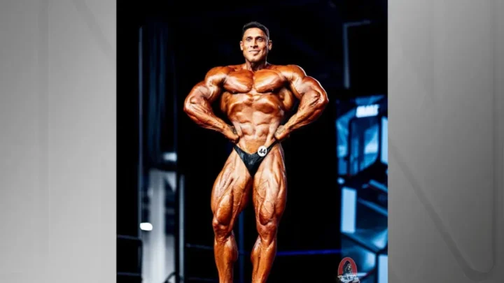Atleta de musculação em posição de pose frontal em palco, exibindo definição muscular sob iluminação de competição, com número de competidor na cintura