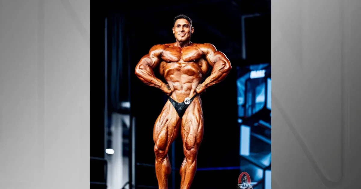Atleta de musculação em posição de pose frontal em palco, exibindo definição muscular sob iluminação de competição, com número de competidor na cintura