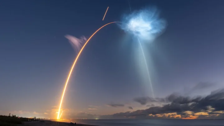 Foto de longa exposição, feita da praia de Cocoa Beach, mostra o arco do Falcon 9 subindo do horizonte até a nuvem azul iluminada pelo sol, com o céu ainda escuro