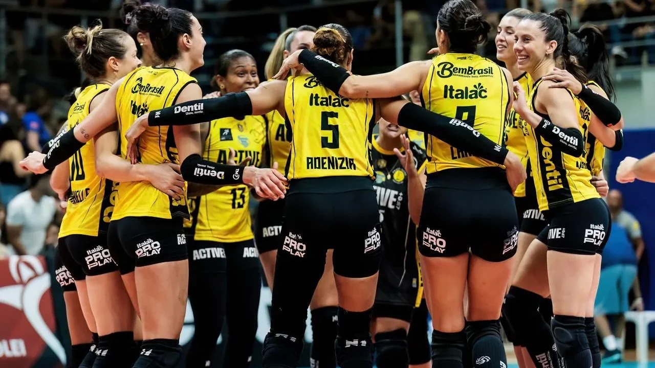 Jogadoras de vôlei com uniforme amarelo e preto, abraçadas em roda, comemorando em uma quadra com torcida ao fundo