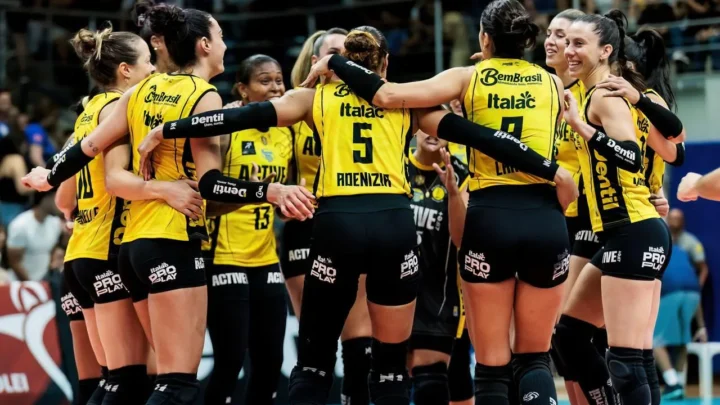 Jogadoras de vôlei com uniforme amarelo e preto, abraçadas em roda, comemorando em uma quadra com torcida ao fundo