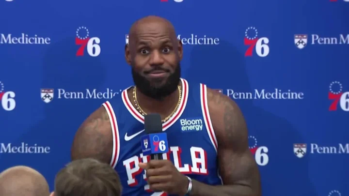 Jogador de basquete em uniforme de time profissional durante entrevista em coletiva de imprensa, com microfone em mão e painel azul com logos de patrocinadores ao fundo