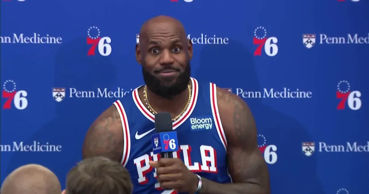 Jogador de basquete em uniforme de time profissional durante entrevista em coletiva de imprensa, com microfone em mão e painel azul com logos de patrocinadores ao fundo