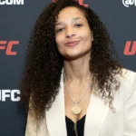Mulher com cabelo comprido ondulado e blazer bege em frente ao painel da UFC durante evento de imprensa