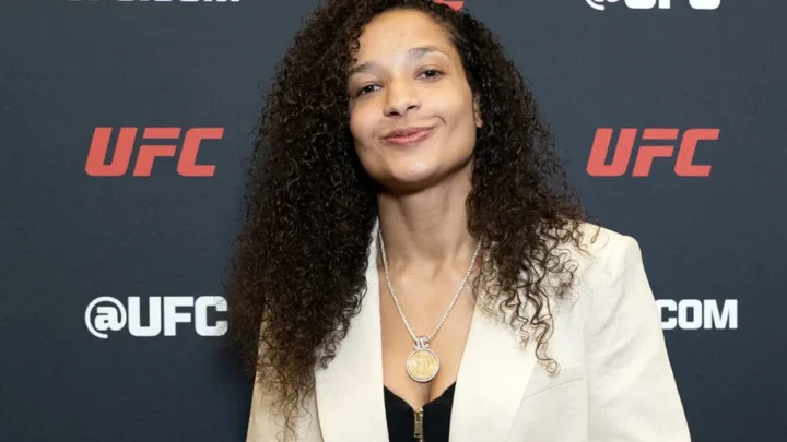 Mulher com cabelo comprido ondulado e blazer bege em frente ao painel da UFC durante evento de imprensa