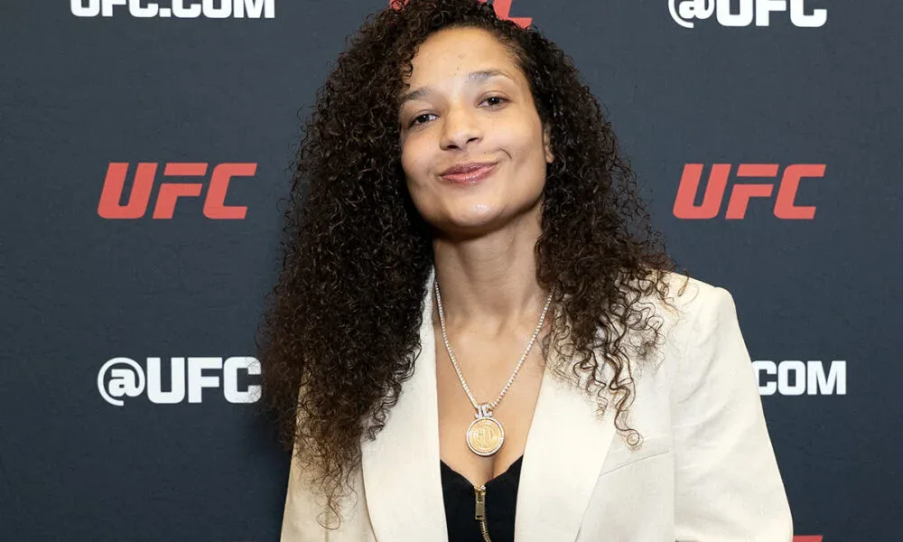 Mulher com cabelo comprido ondulado e blazer bege em frente ao painel da UFC durante evento de imprensa
