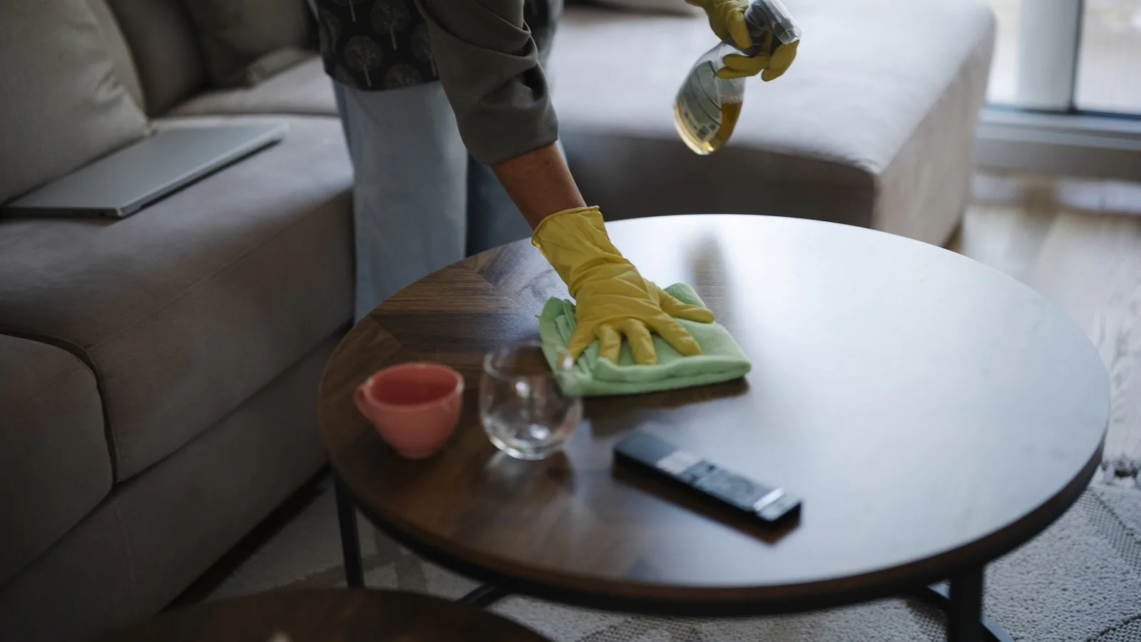Pessoa de luvas amarelas limpa com pano e spray uma mesa de centro numa sala, sem rosto à mostra