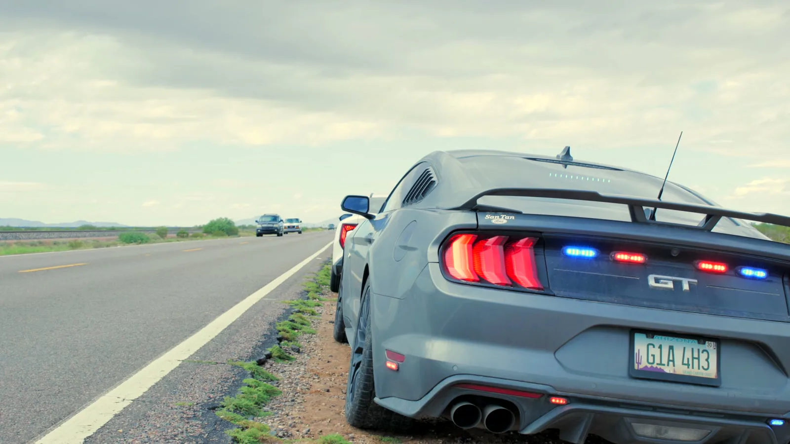 Mustang GT cinza com luzes azuis e vermelhas acesas, parado no acostamento de uma rodovia de pista simples