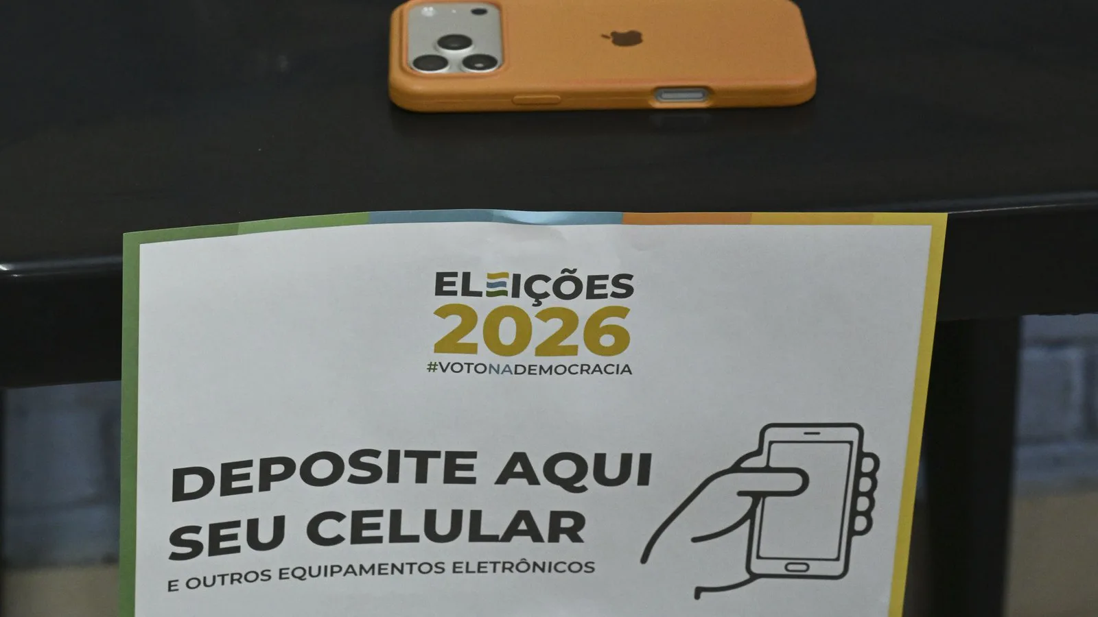 Aviso "Deposite aqui seu celular e outros equipamentos eletrônicos" numa seção eleitoral, com um celular sobre a mesa, no 1º turno, em 4 de outubro de 2026