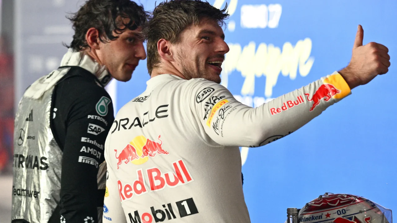 Piloto de macacão branco da Red Bull faz sinal de joinha ao lado de outro piloto de macacão preto da Mercedes, em frente a painel azul de evento