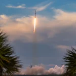 Foguete Falcon 9 sobe sobre a água deixando um rastro de fumaça, emoldurado por palmeiras, no Cabo Canaveral, na Flórida