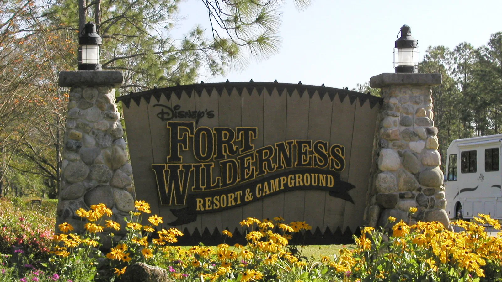Foto de arquivo da placa de pedra e madeira do Disney's Fort Wilderness Resort and Campground, cercada de flores amarelas, com um trailer de camping ao fundo