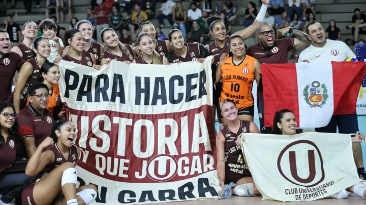 Equipe de voleibol feminino em uniforme vinho comemora vitória segurando bandeiras e faixas com mensagens de apoio em quadra coberta repleta de torcedores