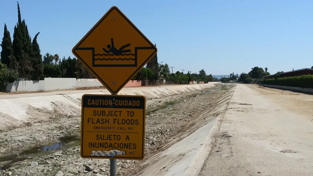 Placa amarela com o desenho de uma pessoa se afogando e o aviso bilíngue "Subject to flash floods / Sujeto a inundaciones" à beira do Pacoima Wash, canal de concreto quase seco em Panorama City, Los Angeles, em 2019 (imagem de arquivo)