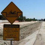 Placa amarela com o desenho de uma pessoa se afogando e o aviso bilíngue "Subject to flash floods / Sujeto a inundaciones" à beira do Pacoima Wash, canal de concreto quase seco em Panorama City, Los Angeles, em 2019 (imagem de arquivo)