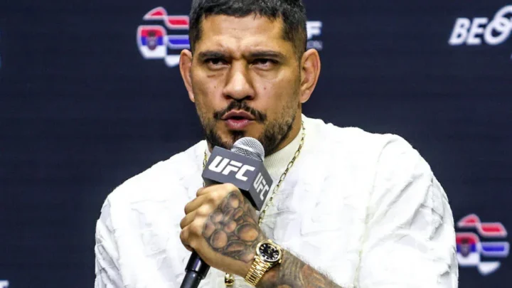 Homem de camiseta branca texturizada, corrente dourada e relógio dourado, segurando microfone da UFC em coletiva com painel escuro ao fundo
