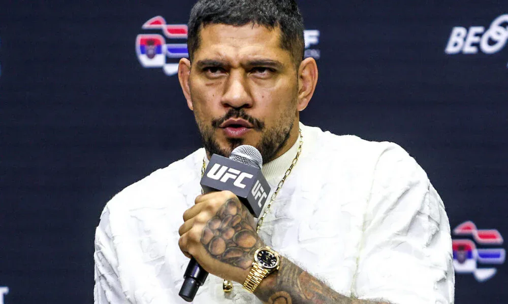 Homem de camiseta branca texturizada, corrente dourada e relógio dourado, segurando microfone da UFC em coletiva com painel escuro ao fundo