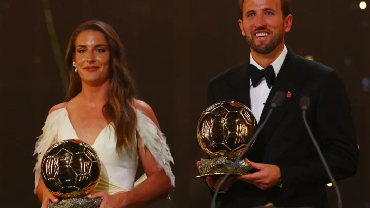 Mulher de vestido branco com penas e homem de smoking seguram troféus de Bola de Ouro em palco escuro