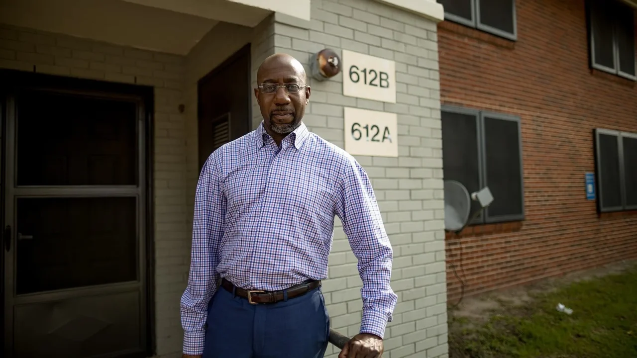 Raphael Warnock, de camisa xadrez, em pé diante de um prédio de tijolos, em foto de agosto de 2020