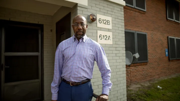 Raphael Warnock, de camisa xadrez, em pé diante de um prédio de tijolos, em foto de agosto de 2020