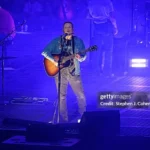 Zach Bryan em show, tocando violão acústico no palco em cenário iluminado com tons de azul e roxo durante apresentação em estádio