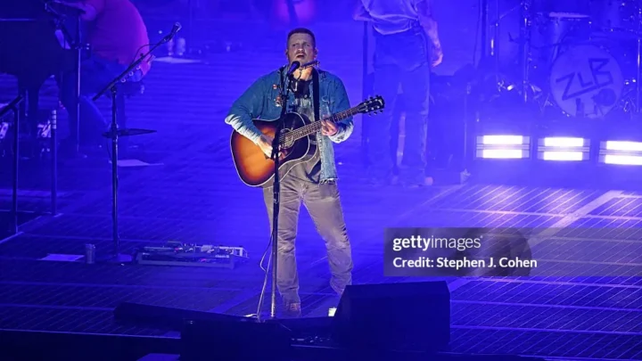 Zach Bryan em show, tocando violão acústico no palco em cenário iluminado com tons de azul e roxo durante apresentação em estádio