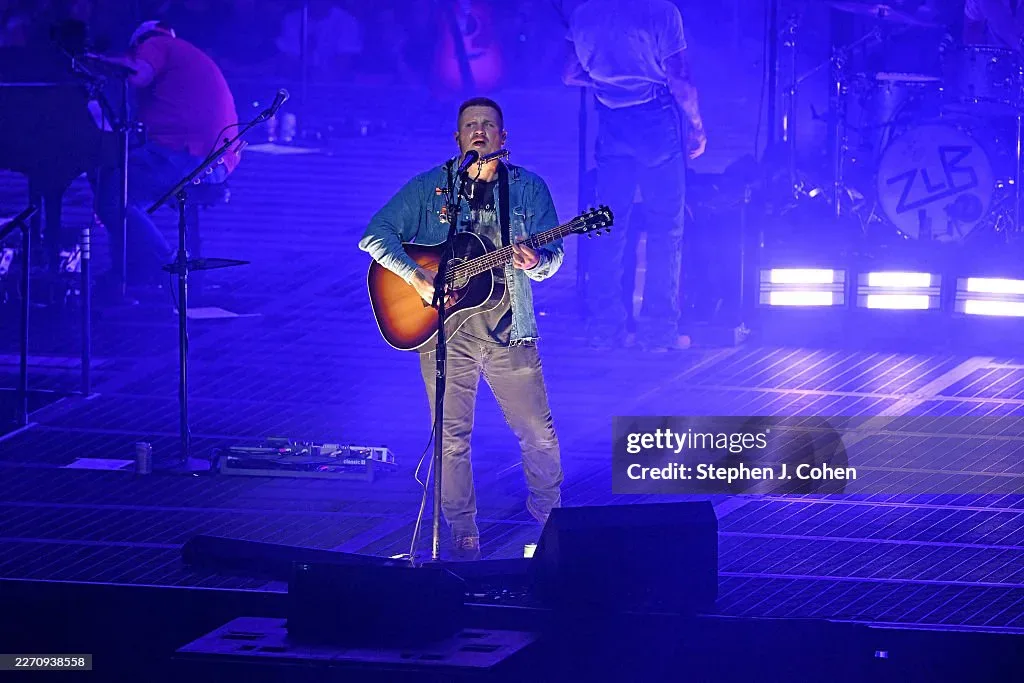 Zach Bryan em show, tocando violão acústico no palco em cenário iluminado com tons de azul e roxo durante apresentação em estádio