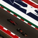 Dois carros de Fórmula 1, um vermelho e um azul escuro, disputam posição em pista de asfalto sobre uma grande estrela branca e vermelha pintada no chão