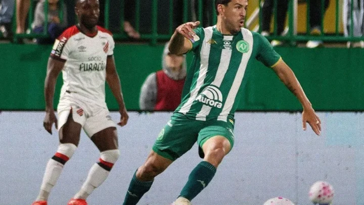 Jogador de camisa verde e listras brancas conduz a bola em campo, enquanto um adversário de branco e vermelho observa ao fundo
